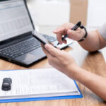 Pessoa consultando a situação documental veicular no celular, com notebook à direita.