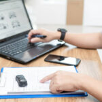 Pessoa digitando no notebook dados de um documento em papel A4, simbolizando atualização cadastral de veículo.