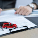 Uma pessoa assinando um documento de transferência de veículo sobre uma mesa, com chave do carro ao lado.