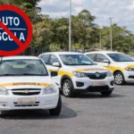 Carros de autoescola modelos Celta, Kwid e Voyage estacionados em rua vazia com placa de proibição ao fundo, representando o fim da obrigatoriedade de fazer autoescola e a ausência de candidatos.