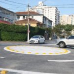 Mini rotatória urbana em cruzamento residencial com carros circulando e sinalização horizontal visível indicando sentido obrigatório