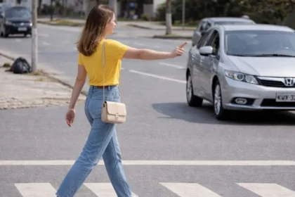 Mulher jovem atravessando a faixa de pedestres em avenida urbana enquanto um carro com motorista aguarda a travessia, demonstrando a prioridade do pedestre no trânsito.