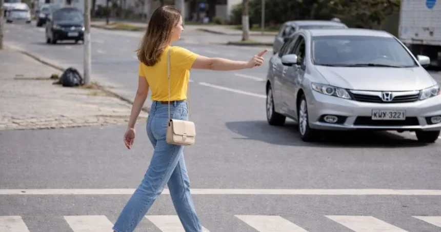 Mulher jovem atravessando a faixa de pedestres em avenida urbana enquanto um carro com motorista aguarda a travessia, demonstrando a prioridade do pedestre no trânsito.