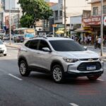 SUV prata com vidros fumê buzinando em uma avenida urbana. Ondas sonoras vermelhas gráficas emanam da grade frontal do veículo sobre o asfalto. Pedestres na calçada e trânsito organizado ao fundo sob luz do dia.